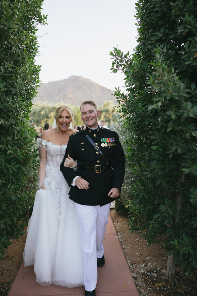 Bride and groom walking on paved path between tall greenery shrubs, smiling.