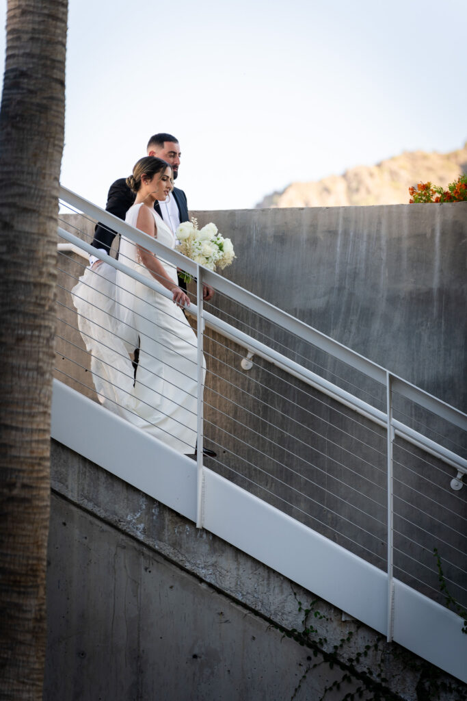 Bride and groom descending stairs outside at Mountain Shadows with groom her holding train of her dress.