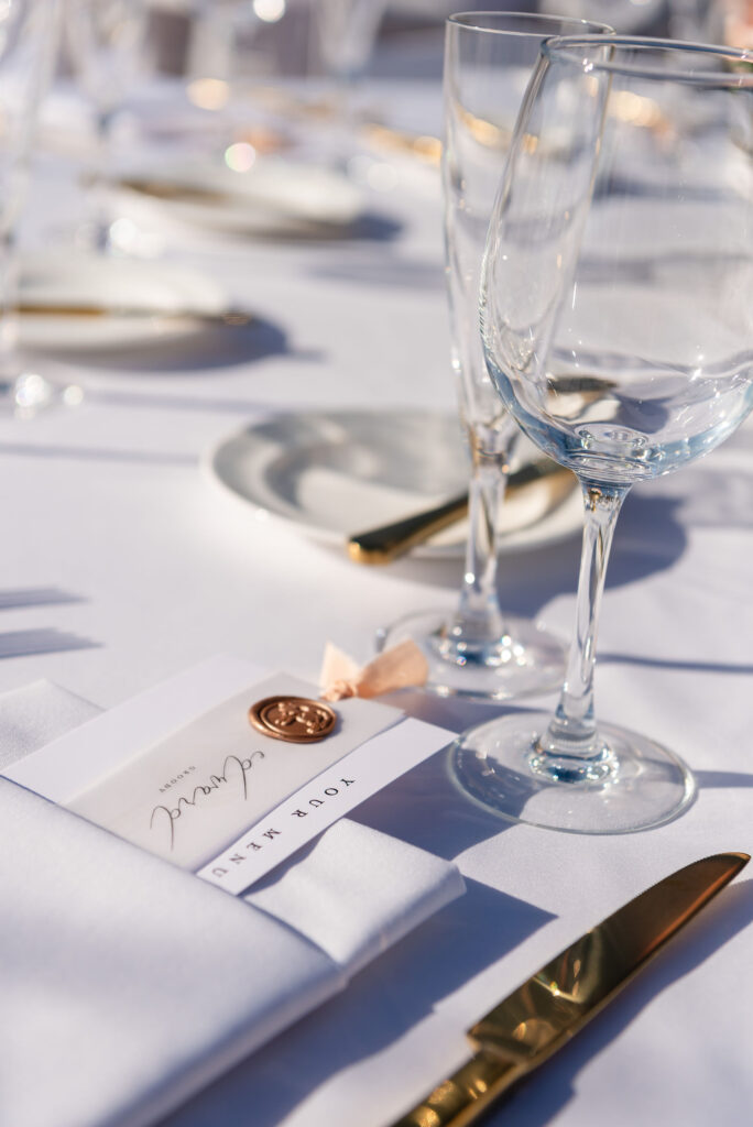 Wedding reception table details of white linen, gold cutlery and white stationary.