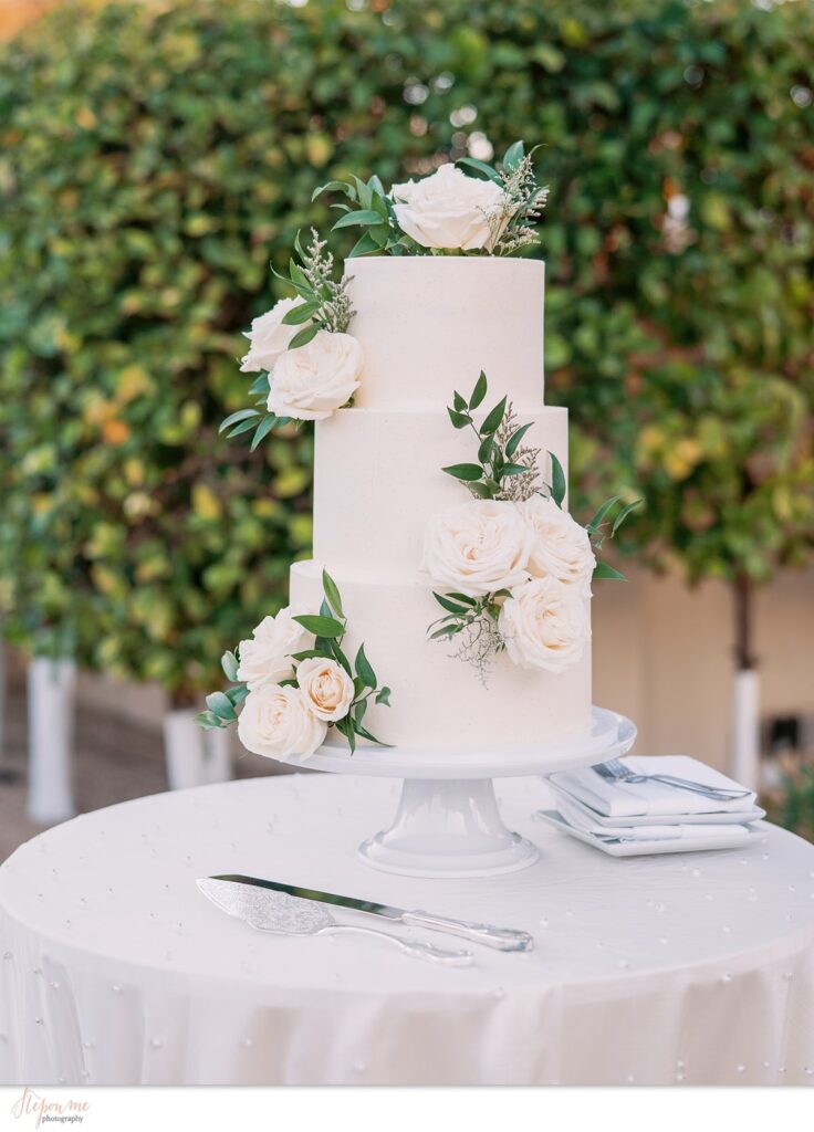 Three tired white wedding cake with flowers and greenery added.