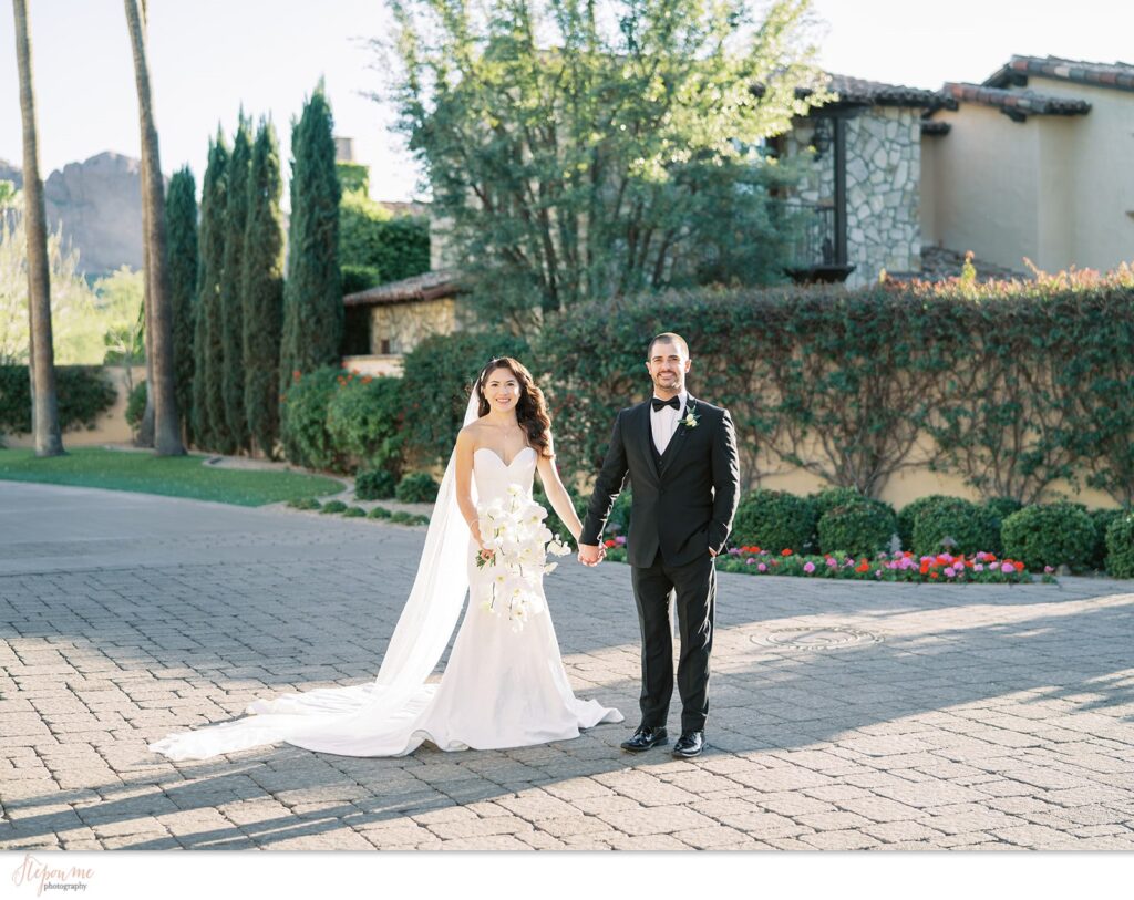 Bride and groom holding hands on brick paved space at Omni resort, smiling at camera.