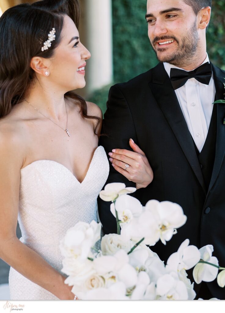 Bride with arm around groom's arm while holding white flower bouquet, both smiling at each other.