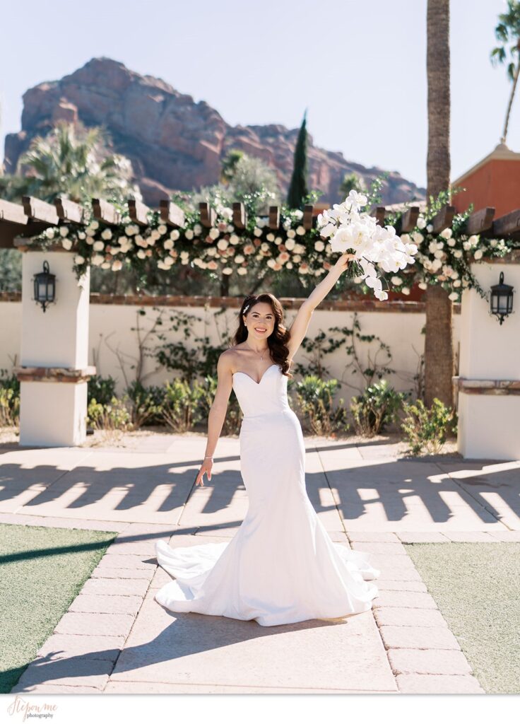 Bride standing in front of Omni resort pergola with Camelback Mountain in background holding bouquet of white flowers up, smiling.