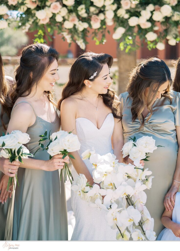 Bride with bridesmaids holding white flowers bouquets, looking down and to side, smiling.