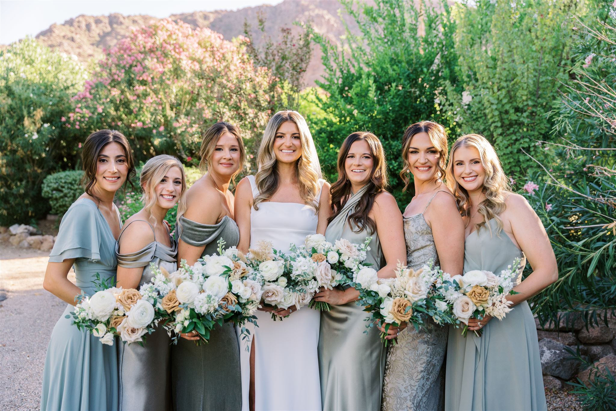 Bride standing with bridesmaids all smiling, in a line all holding bouquets in desert landscape.
