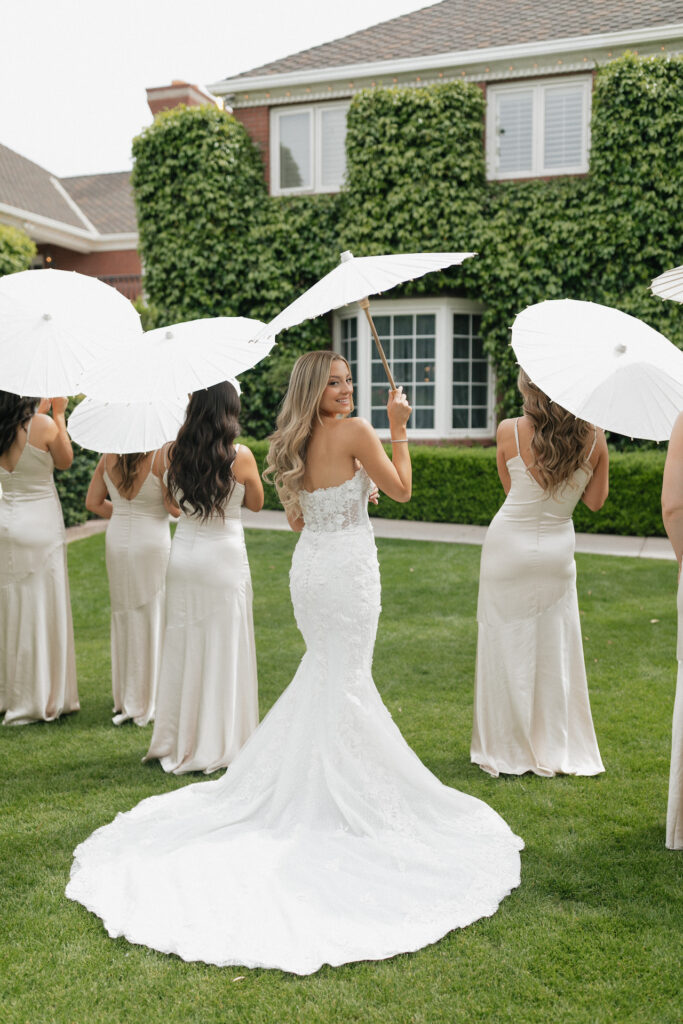 Bride and bridesmaids holding white parasols, bride smiling over should looking behind her.