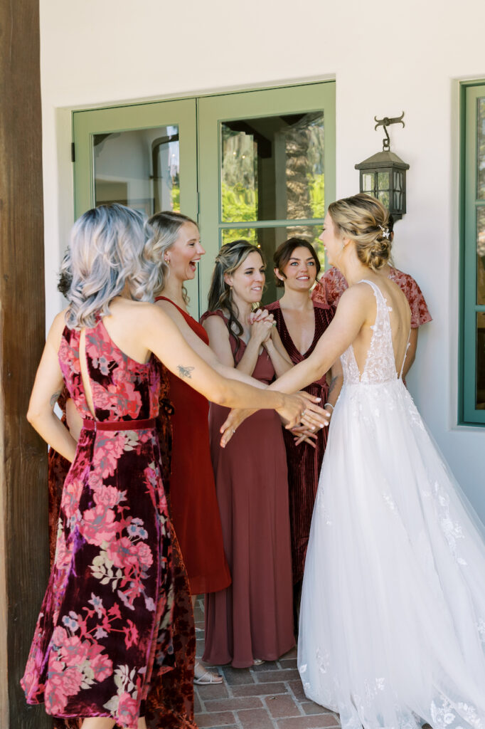 Bridesmaids seeing bride in gown at first look outside double doors of building, all smiling.