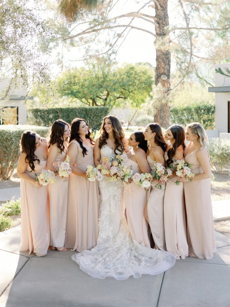 Bride with bridesmaids, standing in a line, all holding bouquets outside, smiling, looking at each other.