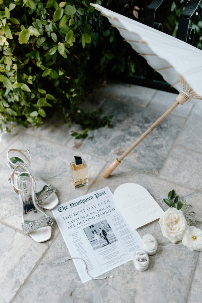 Flat lay of wedding details including white parasol, invitation, rings, jewelry, shoes, and perfume.