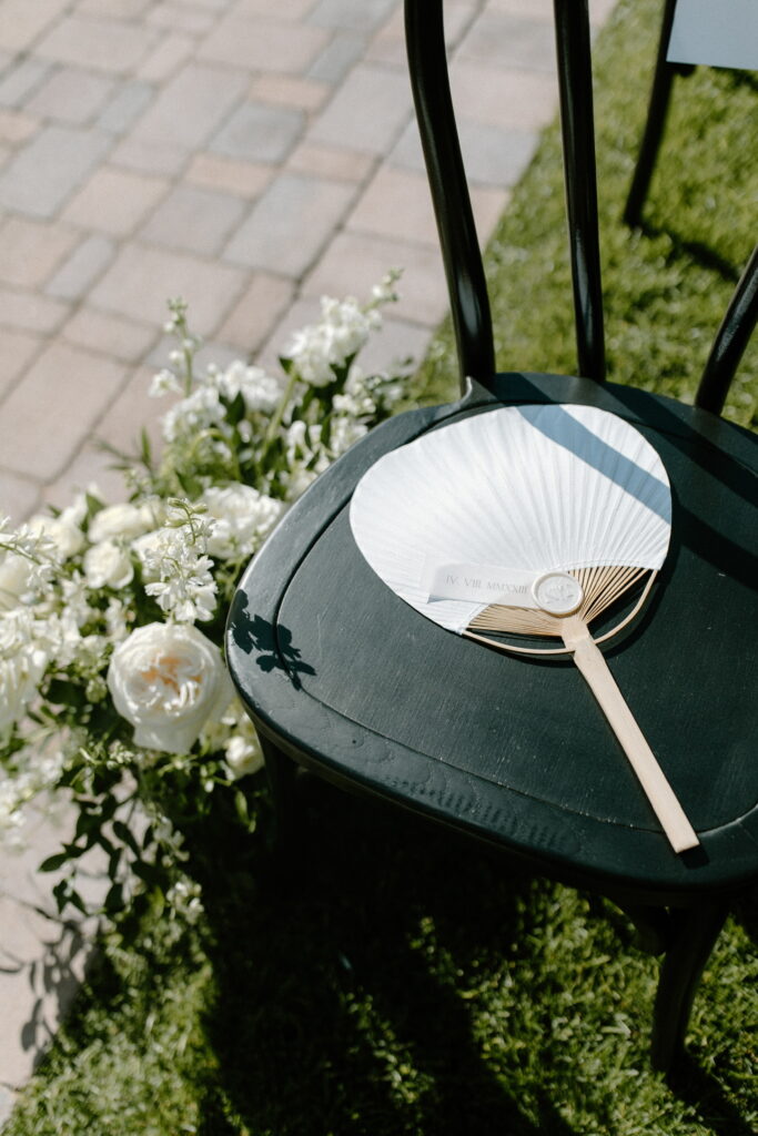 White fan laying on black wedding ceremony chair with floral on ground next to chair and aisle.