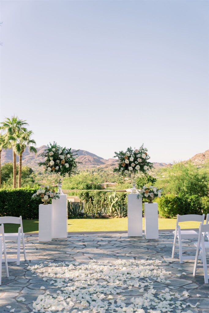 Outdoor wedding ceremony altar space with two difference sized white acrylic pillars with floral arrangements both short and tall on them and rose petals scattered in the aisle.