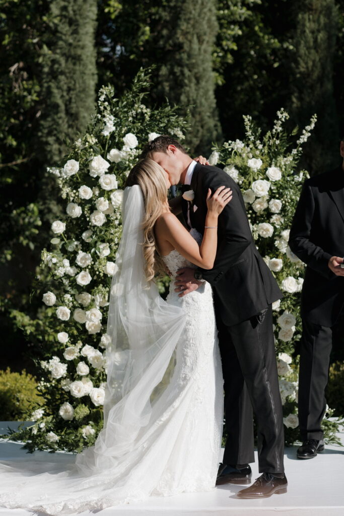 Bride and groom kissing at outdoor wedding ceremony altar space in front of white flowers and greenery floral pillars.