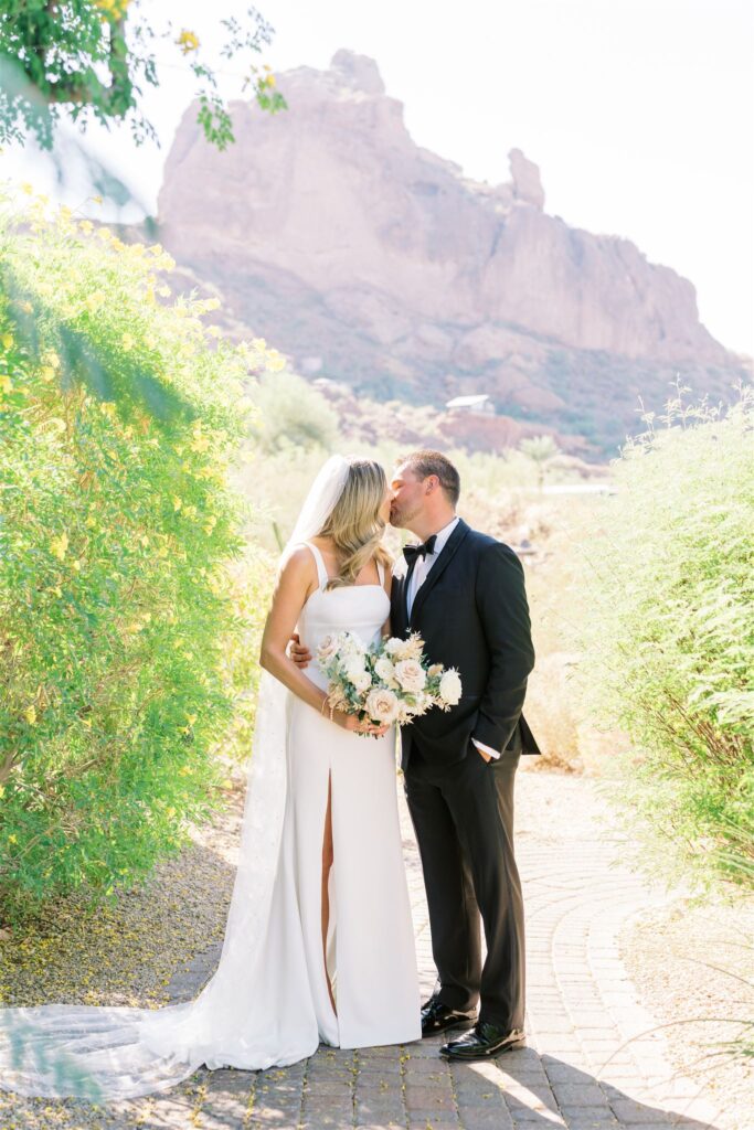 Bride and groom kissing on path in desert landscape with mountain in distance.