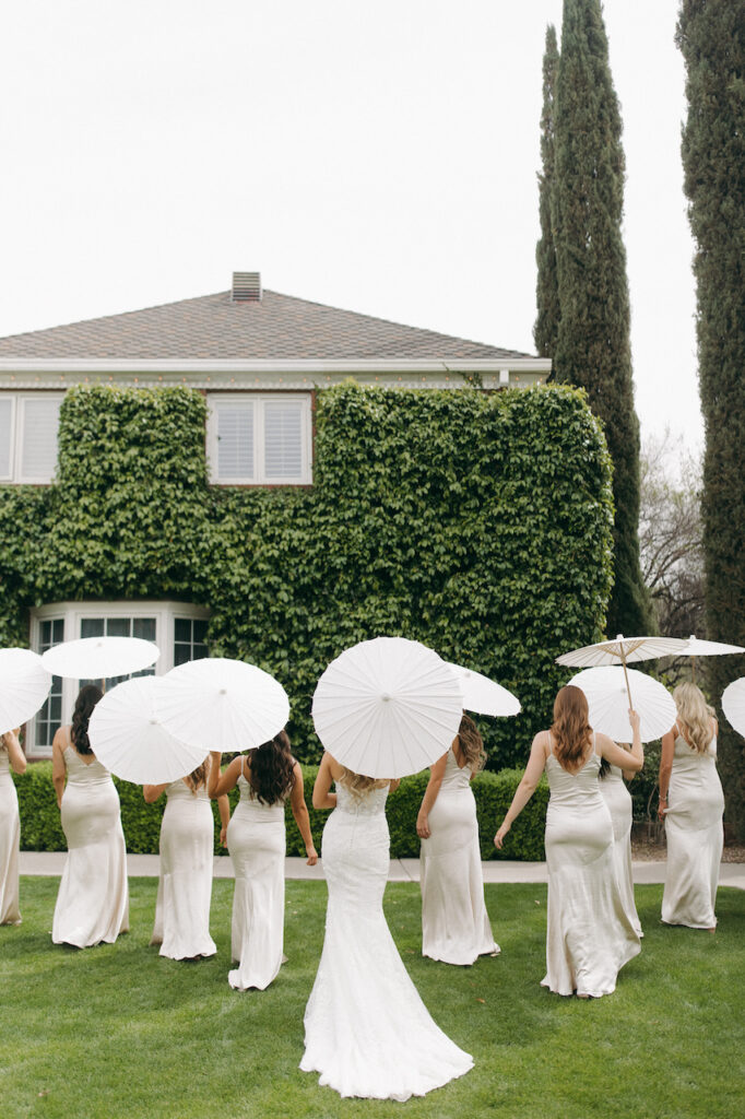 Bride with bridesmaids with their backs all turned holding white parasols in front of building with ivy growing on it.