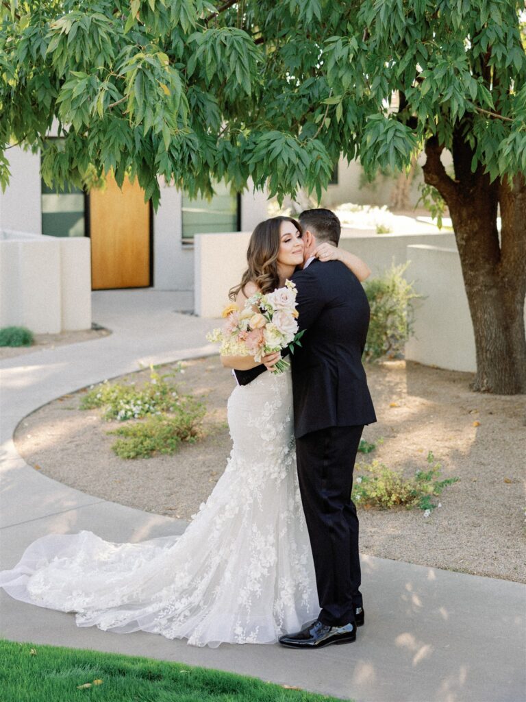 Bride and groom embracing, bride holding bouquet, standing on paved path under tree.