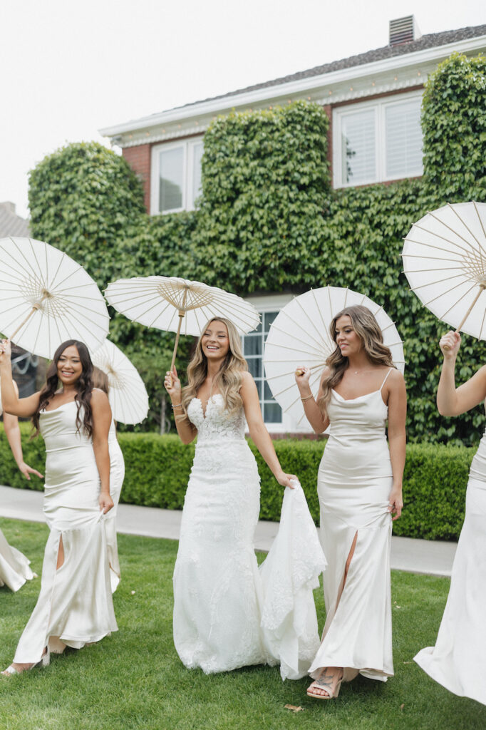 Bride with bridesmaids smiling, walking with white parasols.