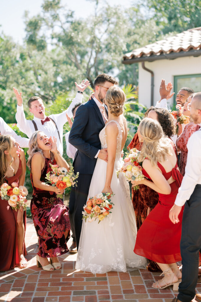Bride and groom kissing in middle of wedding party of groomsmen and bridesmaids celebrating.