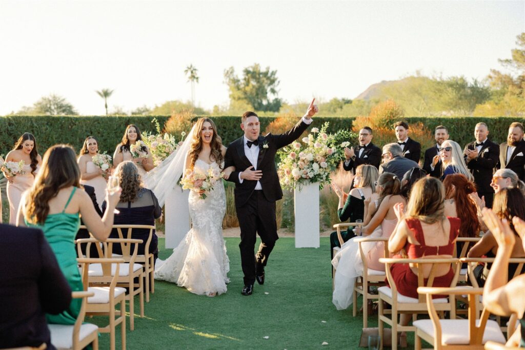 Bride with arm around groom's arm both smiling and walking down wedding ceremony aisle with guests on both side and groom holding one finger up to sky.