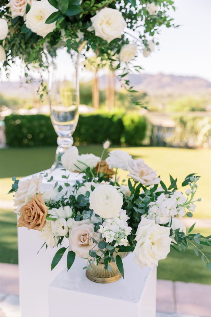 Low floral arrangment in brass vase of white, blush, and gold roses with greenery on a white acrylic pillar.