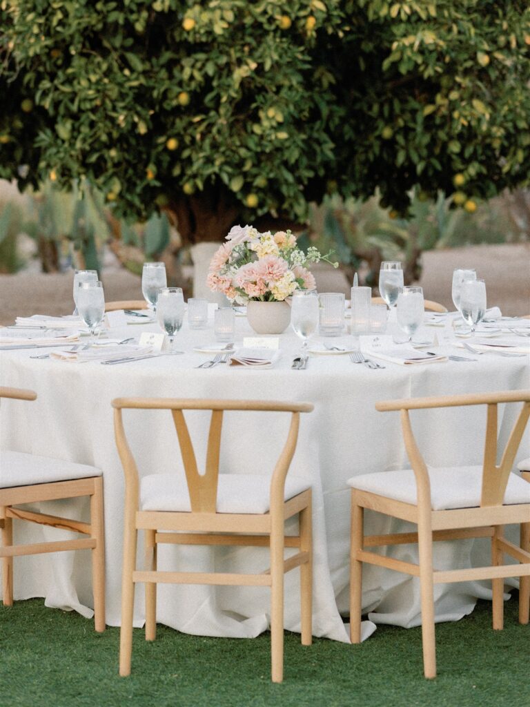 Outdoor wedding reception round table with neutral vase floral centerpiece in middle of set table, white linen, and citrus tree in the background.