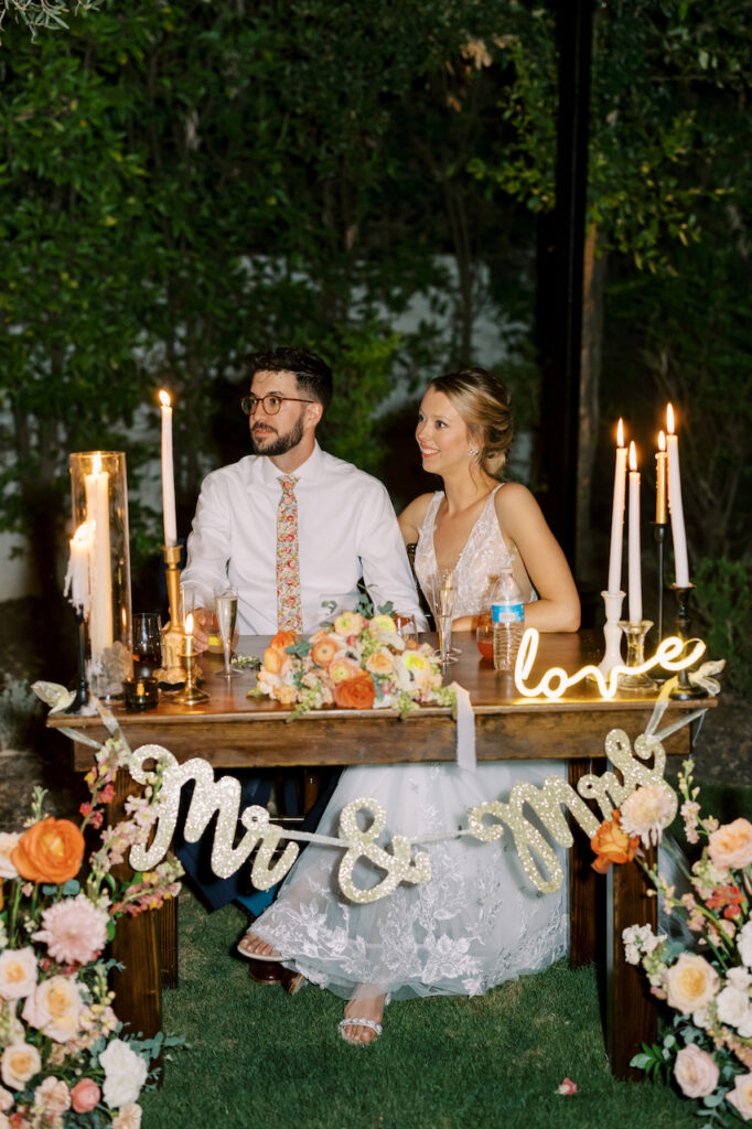 Bride and groom sitting at sweetheart table with a Mr. and Mrs. sign hanging in front, various candles on the table top to the sides and ground floral in front.