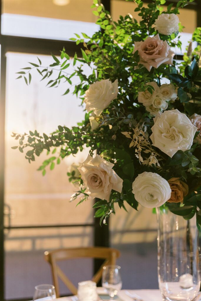 Tall reception centerpiece in glass vase of white, blush, and gold roses with greenery.