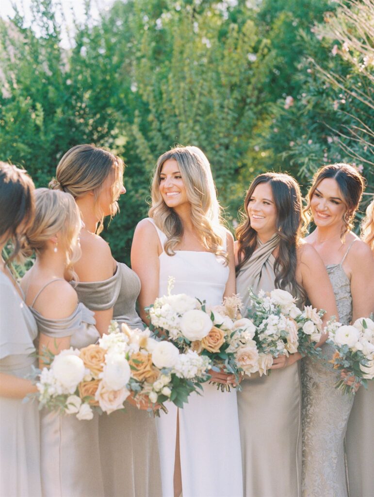 Bride smiling with bridesmaids around her in gray green dresses, all holding bouquets.