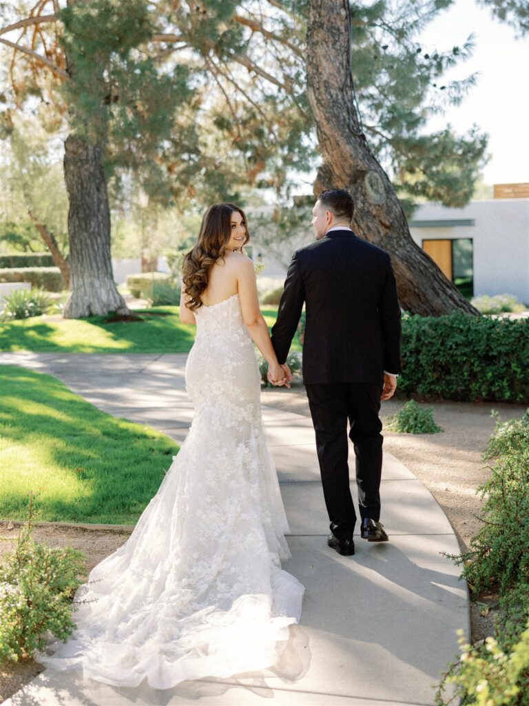 Bride and groom walking down paved path, holding hands and bride is smiling looking back over her shoulder.