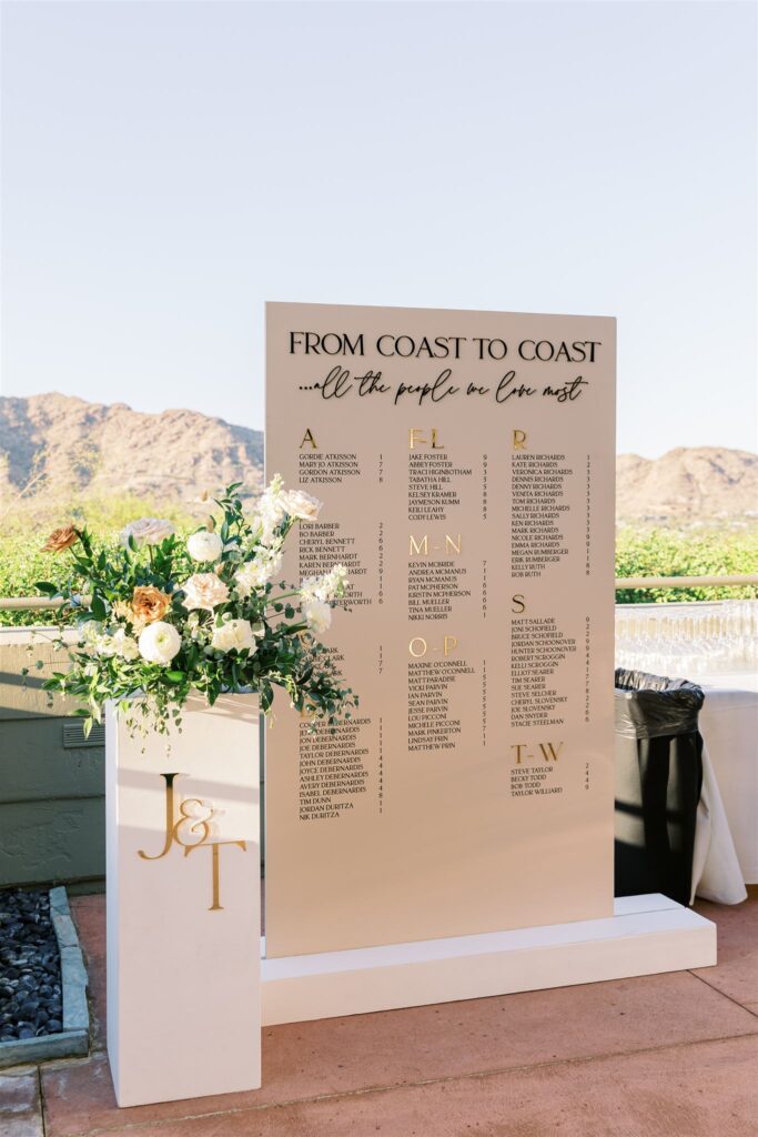 Wedding reception custom escort sign for guests next to floral arrangement on pillar with gold monogram letters.