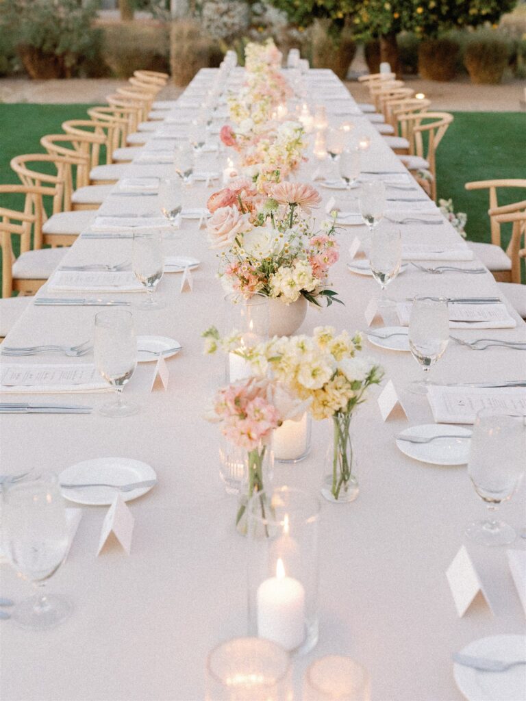 Long reception table with white linen and candles, with centerpieces of flowers in taupe vases and bud vases.