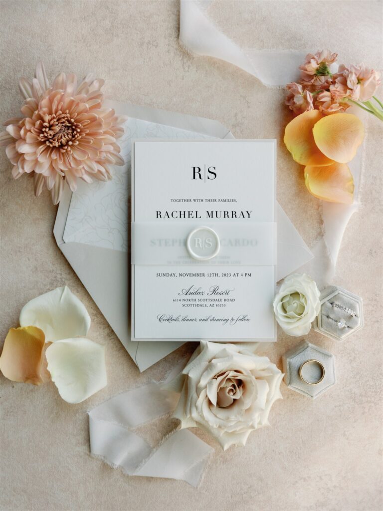 Wedding invitation flat lay of white invitation and taupe envelope with flowers and rings and ribbon around them.