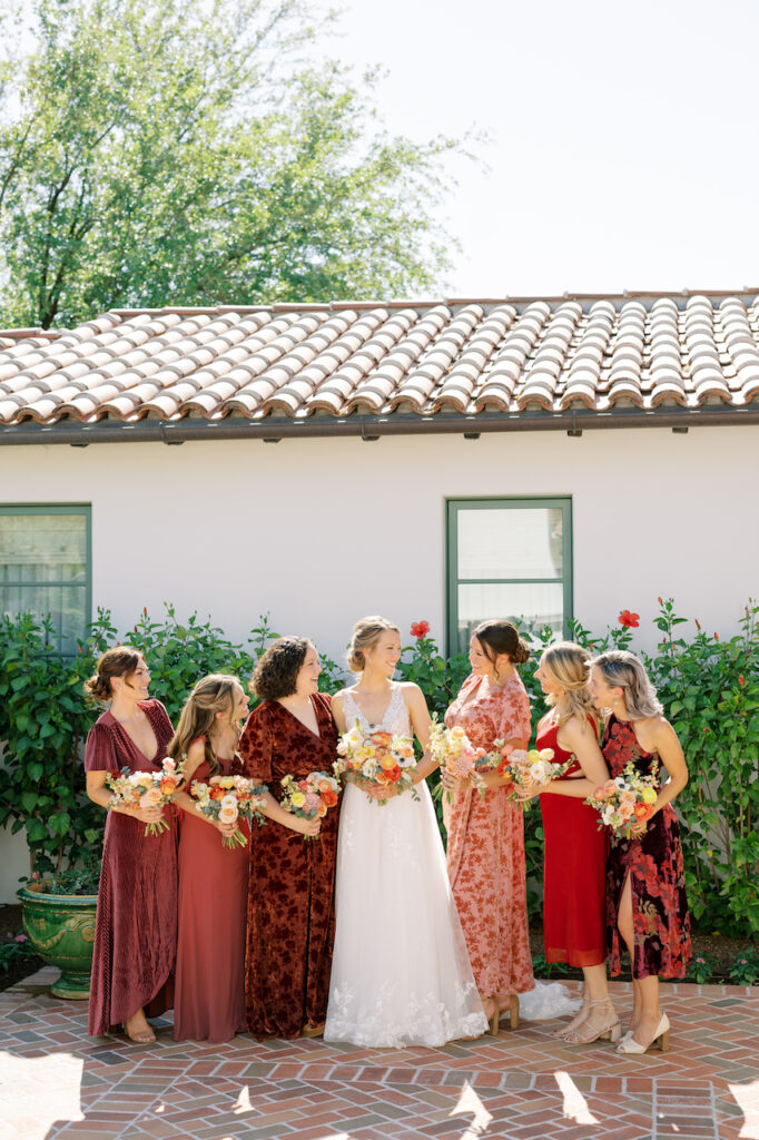 Bride standing in line with bridesmaid in various red gowns all holding bouquets.