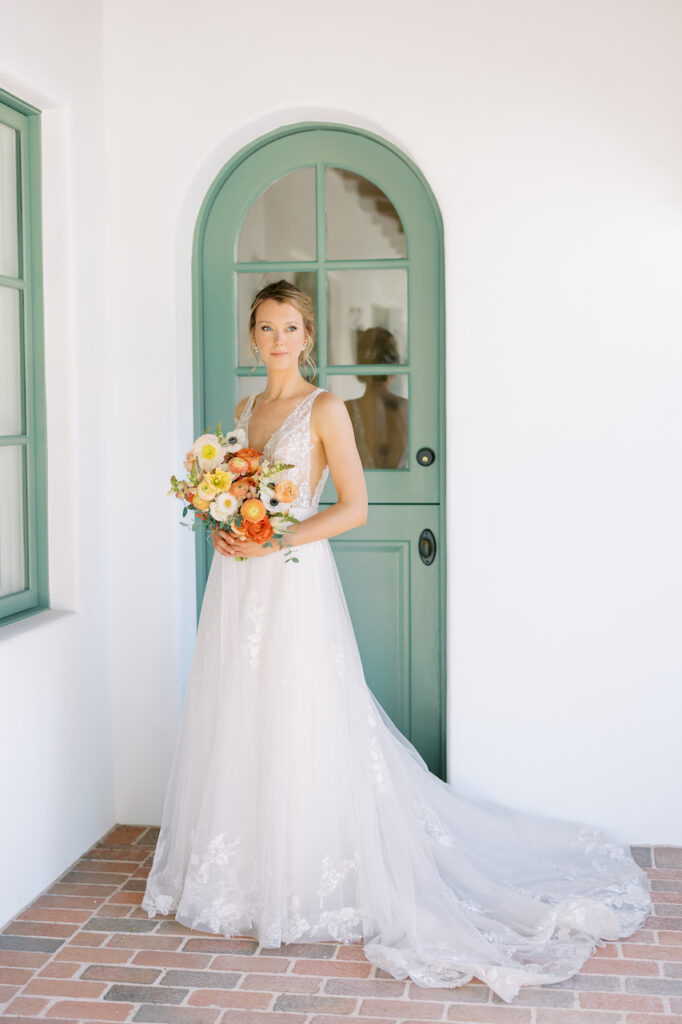 Bride standing in front of green rounded door, holding bouquet.