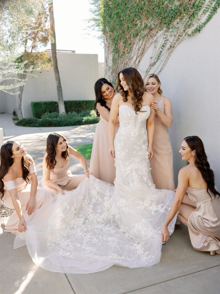 Bride with bridesmaids helping adjust her gown outside on paved area next to building with greenery growing in it.