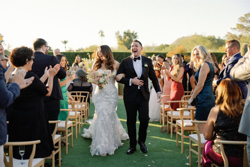 Bride with arm around groom's arm both smiling and walking down wedding ceremony aisle with guests on both side.