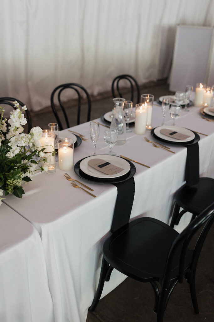 Long wedding reception table with white and black linens and plates with candles and white floral and greenery centerpieces.