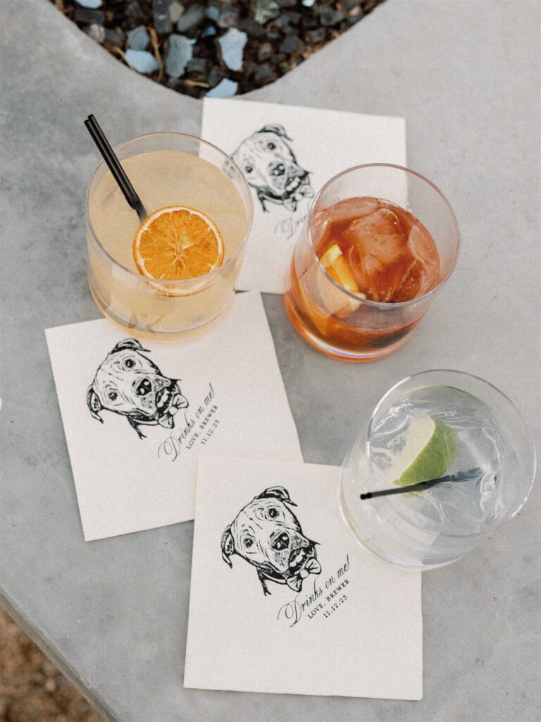 Custom white cocktail napkins with dog image on them and three cocktail drinks with citrus slices in them.