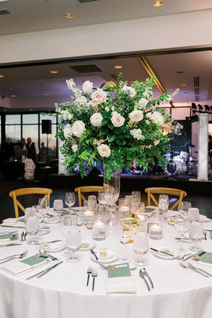 Wedding reception round table with tall floral centerpiece of roses and greenery on tall glass vase.
