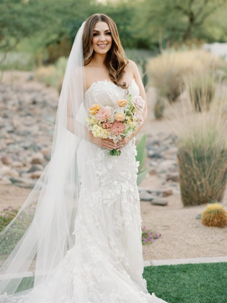 Bride smiling, holding bouquet in gown and with veil and desert background behind her.