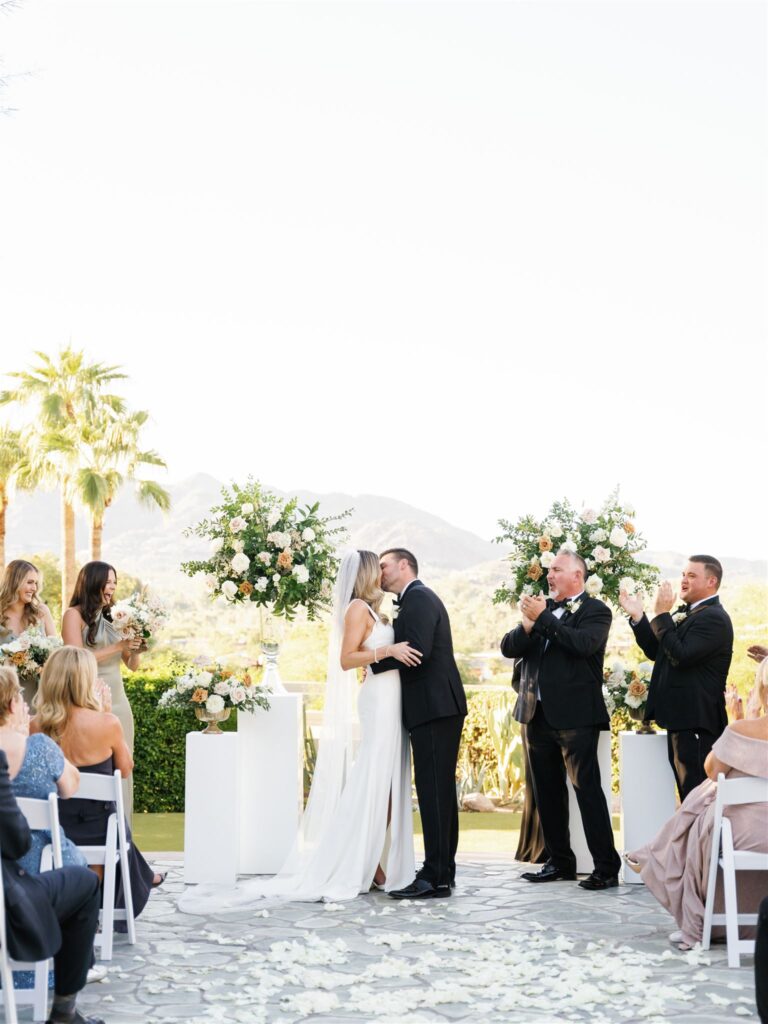 Bride and groom embracing, kissing at outdoor wedding ceremony with floral arrangements on pillars framing them on either side.