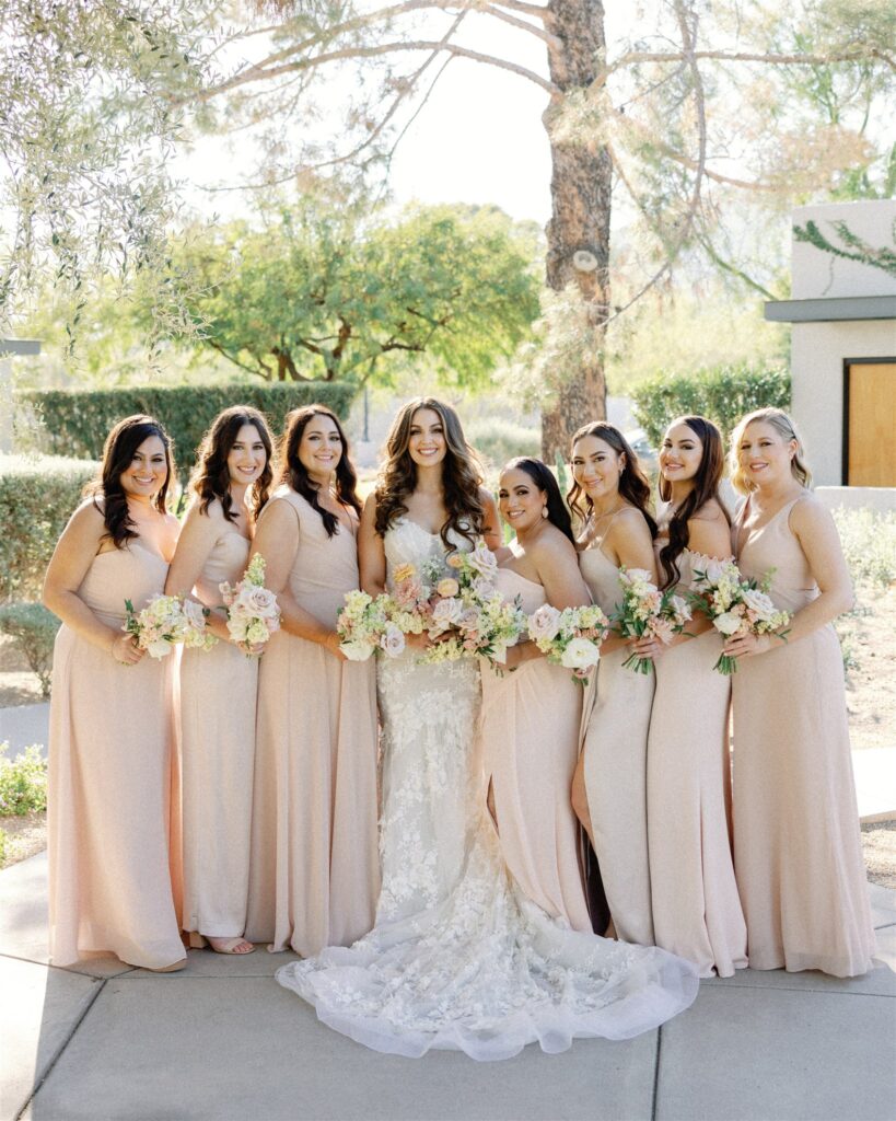 Bride with bridesmaids, standing in a line, all holding bouquets outside, smiling.