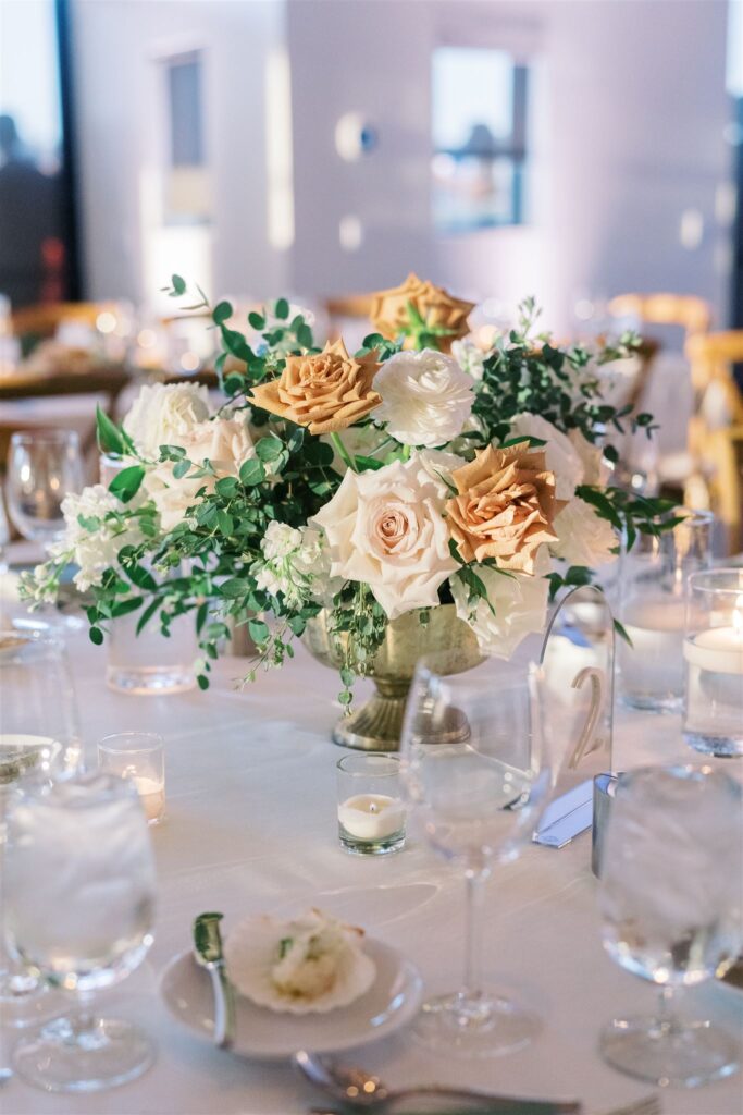 Wedding reception centerpiece of white, blush, and gold roses with greenery in a brass vase.