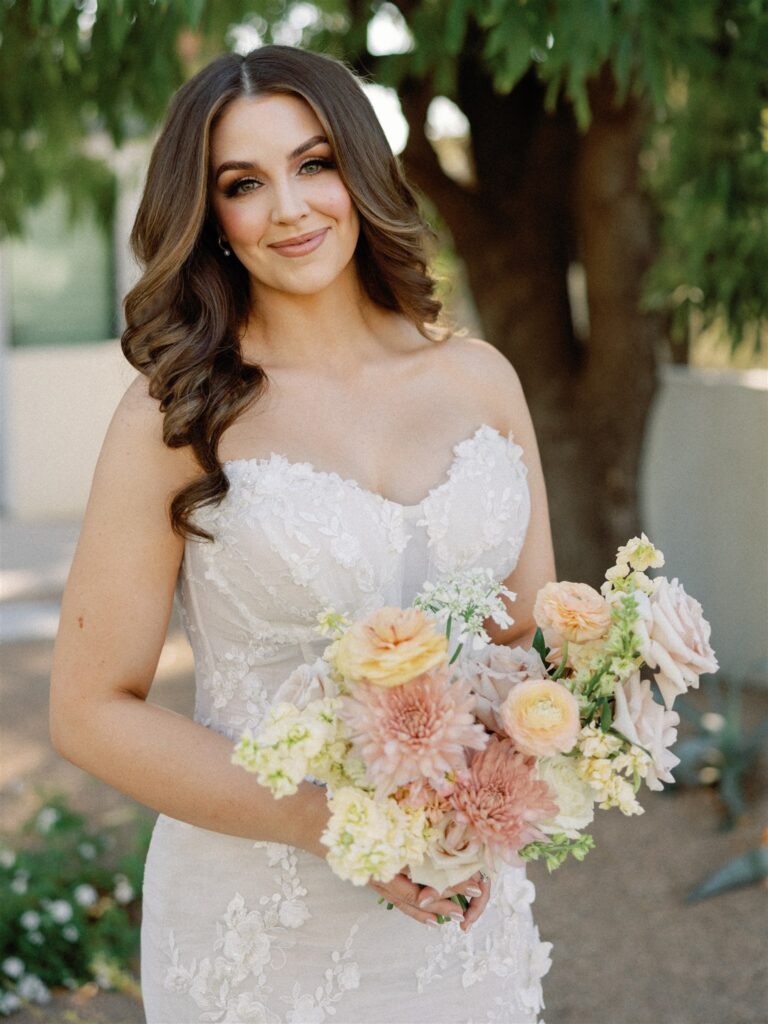 Bride smiling in gown standing in front of tree, holding bouquet of white, pink, peach, and yellow flowers.