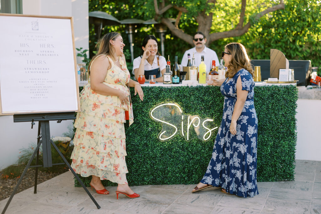 Outdoor wedding reception bar covered in greenery with a neon sign saying "Sips" and two bartenders talking to two guests.