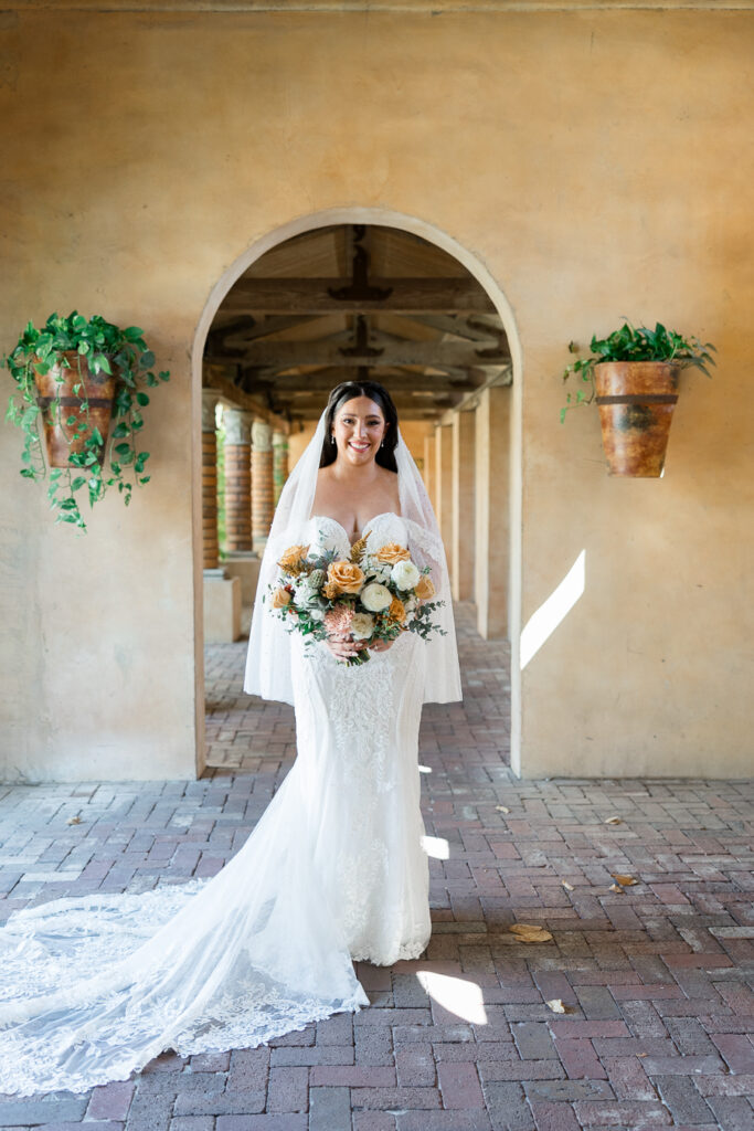 Bride standing in building archway holding bouquet of gold and white flowers.