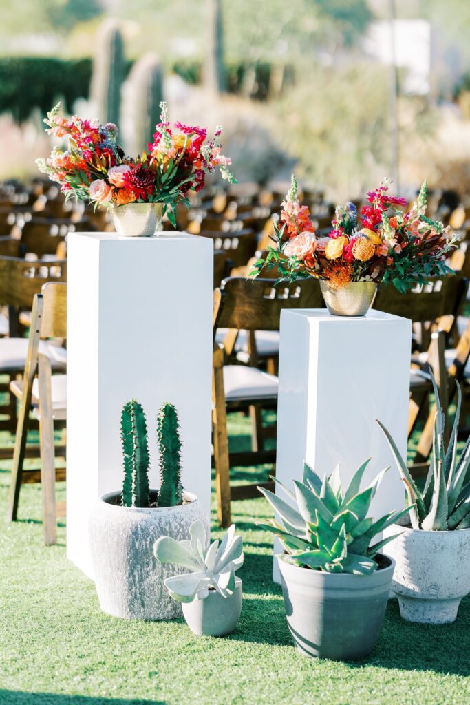 Back of wedding ceremony aisle floral decor of arrangements on white acrylic pillars and desert plants in pots on ground.