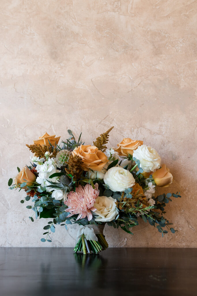 Bridal bouquet placed on table and leaning against a wall with flowers in colors of gold, white, pink, and greenery.