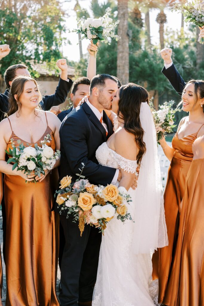 Bride and groom kissing with their wedding party celebrating around them.