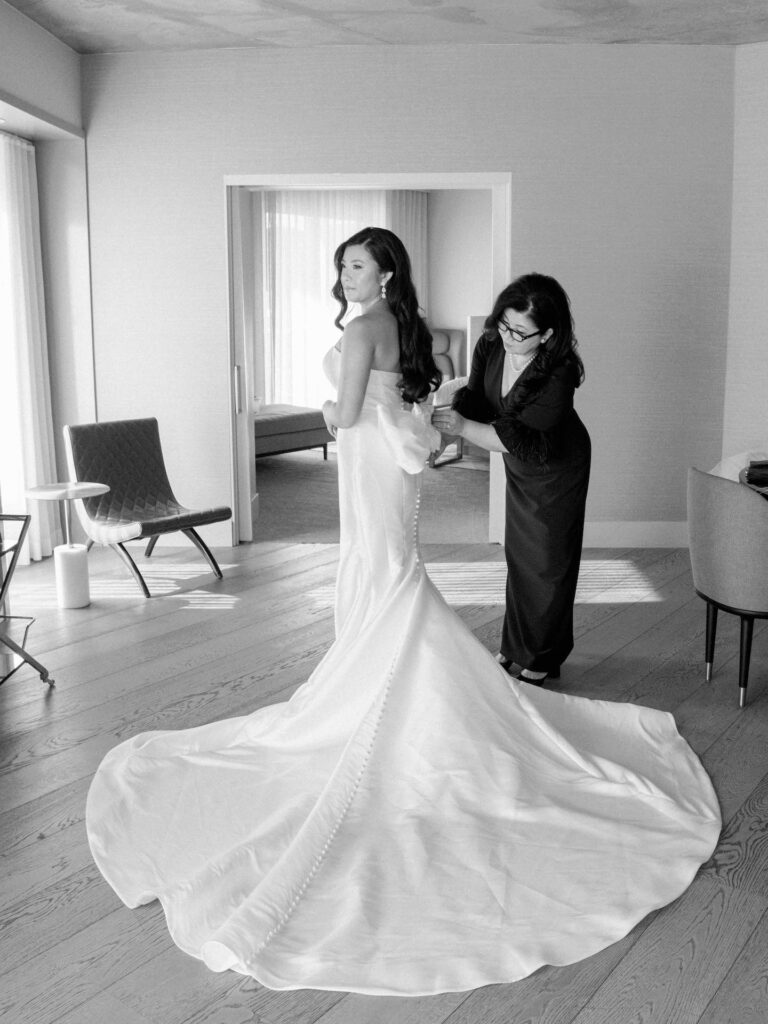 Bride receiving help with buttoning the back of her gown from another woman, her dress train spread out large on the ground.