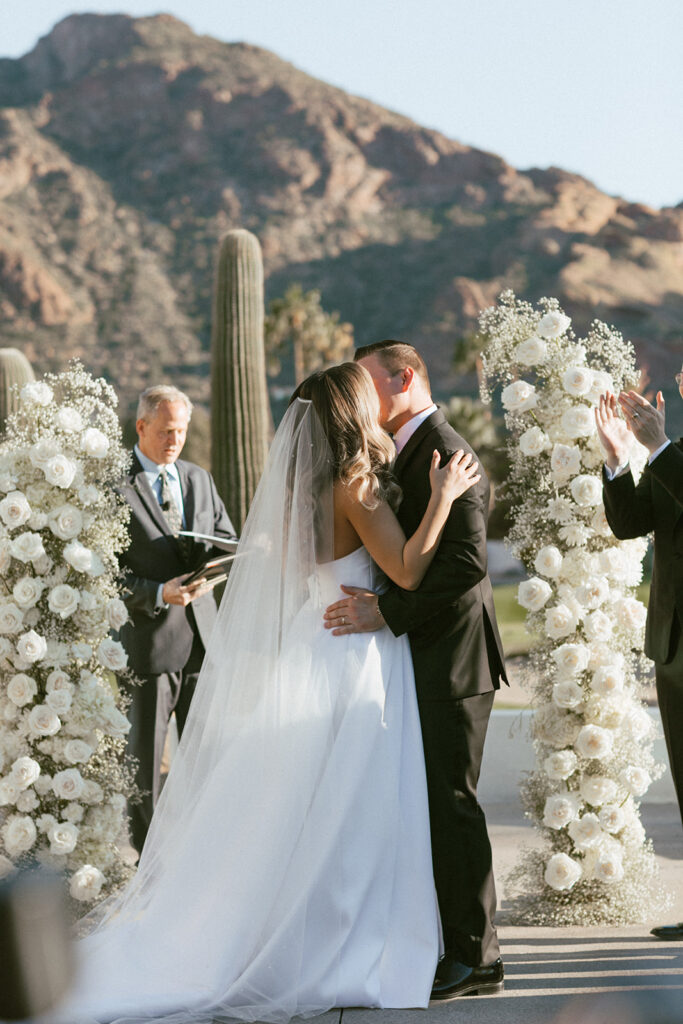 Bride and groom kissing in wedding ceremony altar space with white floral columns on either side of them.